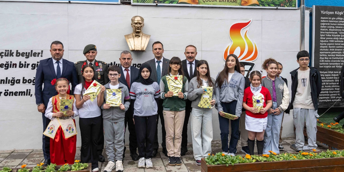 Atatürk büstü ve meşale figürünün önünde, aralarında resmi kıyafetli yetkililerin ve çocukların bulunduğu bir grup, ellerinde hediye paketleriyle yan yana duruyor. Çocuklardan biri geleneksel kıyafetler giyerken, diğerleri günlük kıyafetleriyle yer alıyor. Arka planda duvarda Atatürk'ün sözlerinin yer aldığı bir pano ve sağ tarafta 