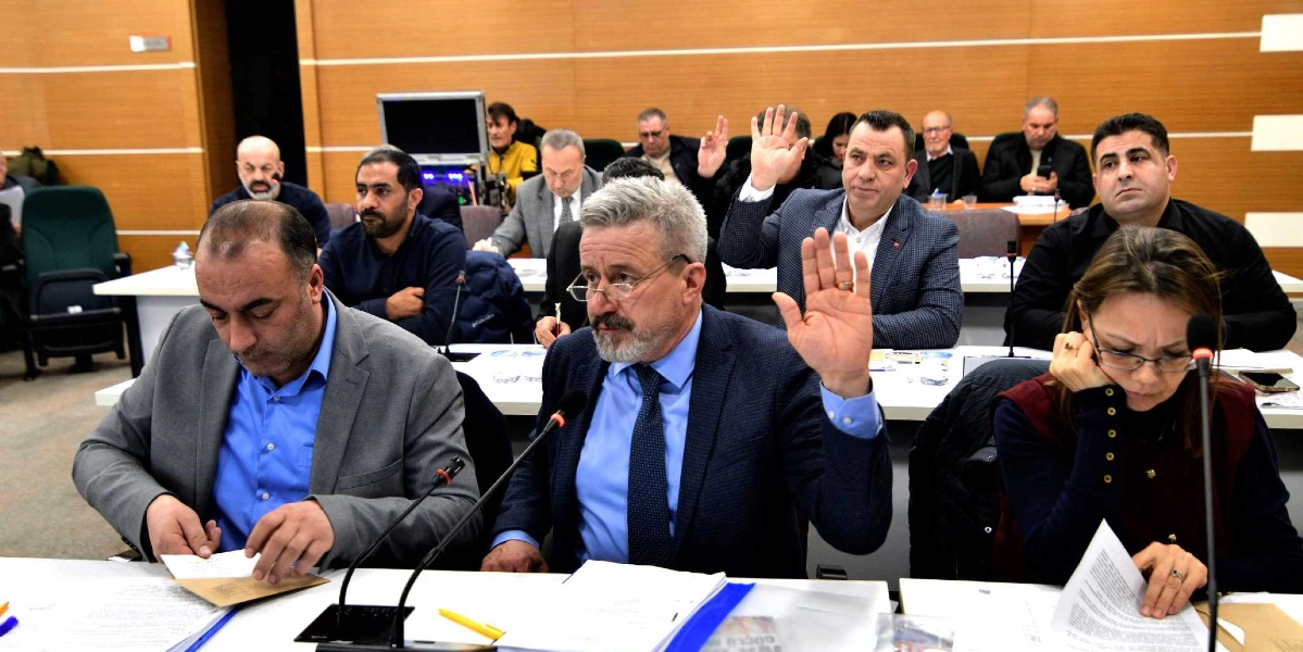 Resmi bir toplantı salonunda, masada oturan bir grup insan oylama yapıyor. Ön planda, gri ceketli ve mavi gömlekli bir adam elini kaldırarak oy kullanıyor; yanında ise evraklarıyla ilgilenen başka bir kişi ve önündeki kağıtları inceleyen gözlüklü bir kadın bulunuyor. Arka planda ise toplantıya katılan diğer üyeler oturuyor ve bazıları da el kaldırarak oylamaya katılıyor.