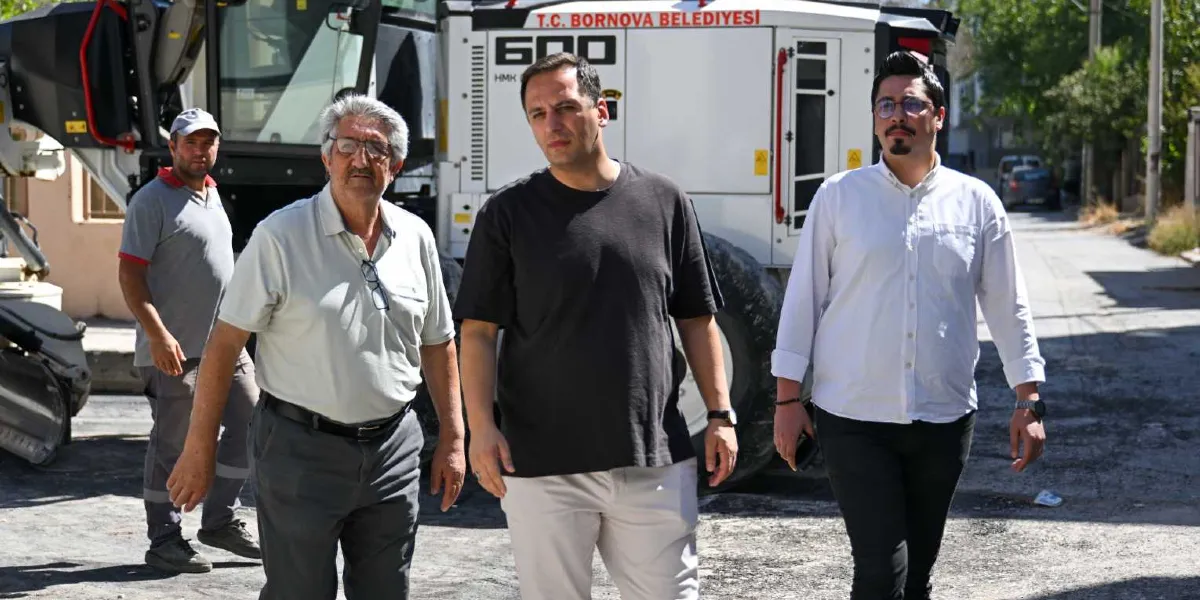 Bornova Belediyesi'ne ait bir iş makinesinin önünde yürüyen üç erkek; gri polo yaka tişörtlü bir çalışan, beyaz gömlekli ve gözlüklü bir kişi ile siyah tişörtlü bir yetkili, devam eden yol çalışmalarını incelemek üzere bir sokakta ilerliyor.