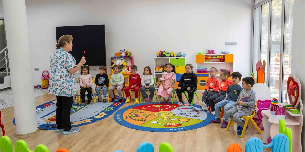 Kreş ortamında, üzerinde desenli bir önlük bulunan bir eğitmen, elinde tuttuğu diş fırçasıyla çocuklara diş sağlığı eğitimi veriyor. Eğitmenin karşısında yarım daire şeklinde dizilmiş sandalyelerde oturan yaklaşık on çocuk, dikkatle eğitmeni dinliyor. Sınıfın zemininde üzerinde harfler ve hayvan figürleri bulunan renkli oyun halıları yer alırken, arka planda oyuncak rafları ve çeşitli çocuk oyuncakları bulunuyor.