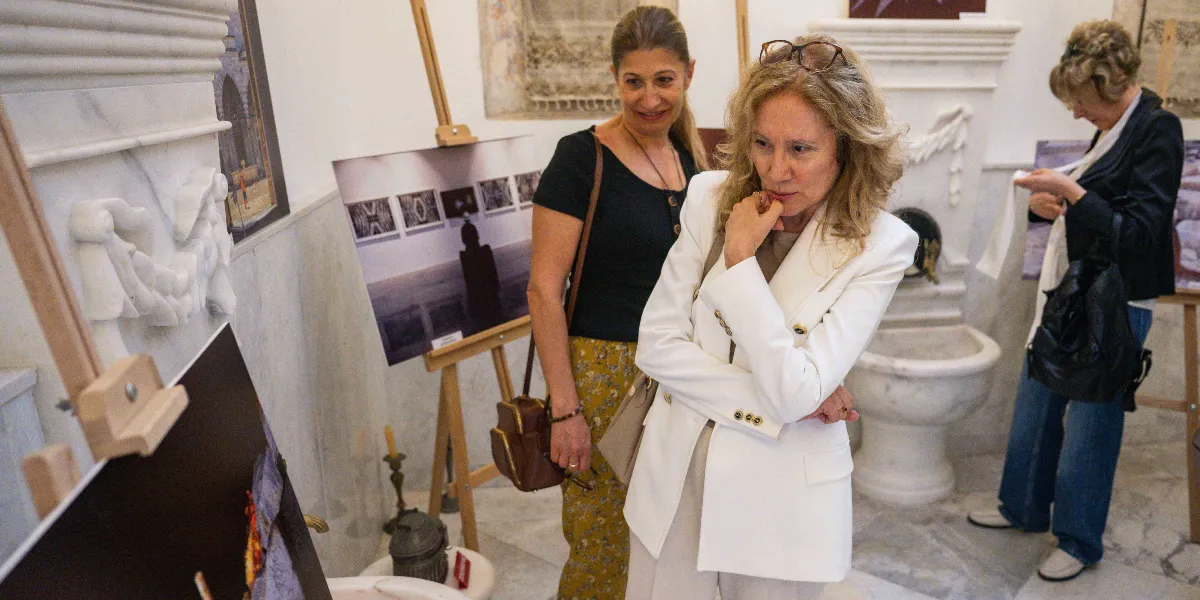 Tarihi bir mekanda düzenlenen sergide, şövale üzerinde sergilenen fotoğrafları inceleyen üç kadın bulunuyor. Ön planda beyaz ceketli, sarı saçlı bir kadın elini çenesine koymuş dikkatle bir fotoğrafa bakarken, yanında siyah tişörtlü ve desenli pantolonlu bir kadın ona eşlik ediyor. Arka planda ise siyah ceketli ve kot pantolonlu başka bir kadın elindeki bir belgeyi inceliyor. Mekanda mermer detaylar ve klasik mimari unsurlar göze çarpıyor.