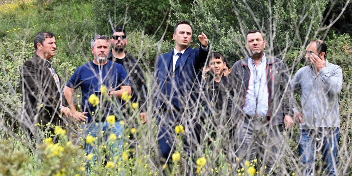 Lacivert takım elbiseli bir erkek, yeşil ve ağaçlık bir alanda yanındaki gruba eliyle bir yönü işaret ederek bir şeyler anlatıyor. Grubun geri kalanında farklı yaşlarda erkekler bulunuyor; bazıları günlük kıyafetlerle, bazıları ise ceketle eşlik ediyor. Ön planda sarı çiçekli çalılar ve kuru dallar yer alıyor.