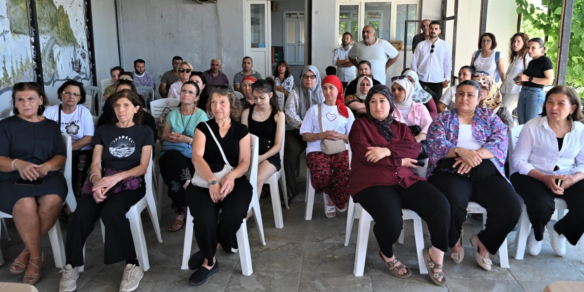 Yarı açık bir alanda beyaz plastik sandalyelerde oturarak bir sunumu veya etkinliği dinleyen, çoğunluğu kadınlardan oluşan kalabalık bir grup. Katılımcılar gündelik kıyafetler içinde, dikkatli bir şekilde karşıya bakıyorlar. Arka planda duvarın bir kısmında ağaç ve doğa temalı bir duvar resmi bulunuyor, yan tarafta ise bir bina girişi ve ayakta duran birkaç erkek katılımcı yer alıyor.