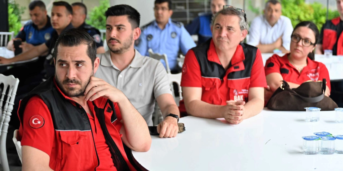 Bir toplantı salonunda beyaz masaların etrafında oturan, aralarında kırmızı yelekli arama kurtarma görevlilerinin ve polis memurlarının bulunduğu bir grup insan dikkatle bir noktaya bakıyor. Ön planda, üzerinde Türk bayrağı arması bulunan kırmızı yelekli bir erkek, elini çenesine dayamış şekilde dinliyor; yanında ise yine kırmızı yelekli bir erkek ve gözlüklü bir kadın oturuyor. Arka planda ise mavi üniformalı polisler ve diğer katılımcılar yer alıyor.