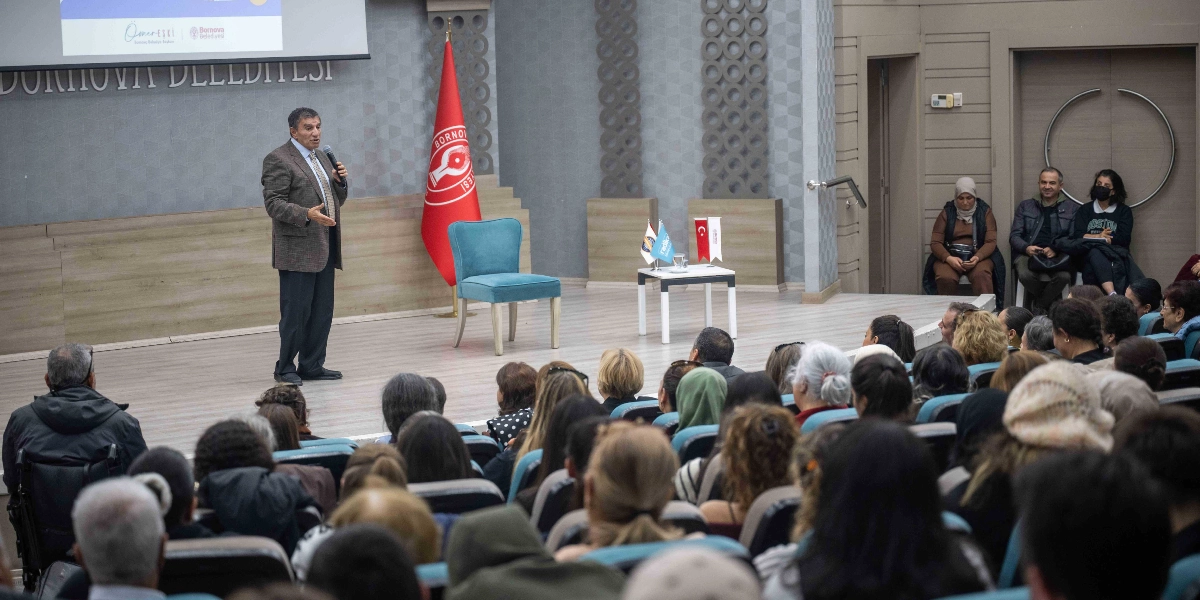 Bornova Belediyesi'ne ait bir konferans salonunda, kürsüde elinde mikrofonla konuşma yapan orta yaşlı bir erkek ve onu dinleyen kalabalık bir izleyici grubu. Konuşmacının arkasında Bornova Belediyesi'nin kırmızı bayrağı, yanında ise üzerinde küçük Türk bayrakları bulunan beyaz bir sehpa ve mavi bir sandalye yer alıyor. Salonun arka kısmında ise izleyiciler oturarak sunumu takip ediyor.