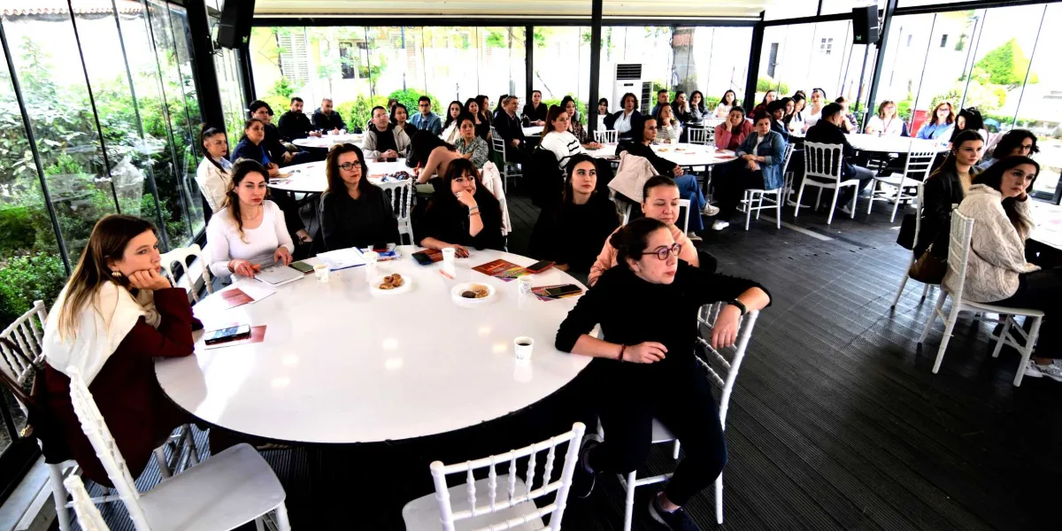 Camla kaplı, aydınlık bir salonda yuvarlak masalar etrafında toplanmış, çoğunluğu kadınlardan oluşan bir grup katılımcı, dikkatle bir sunumu veya konuşmacıyı dinliyor. Masaların üzerinde not defterleri, kalemler ve küçük atıştırmalık tabakları bulunuyor. Katılımcılar gündelik kıyafetler içinde, birbirine dönük veya sahneye odaklanmış şekilde oturuyor.