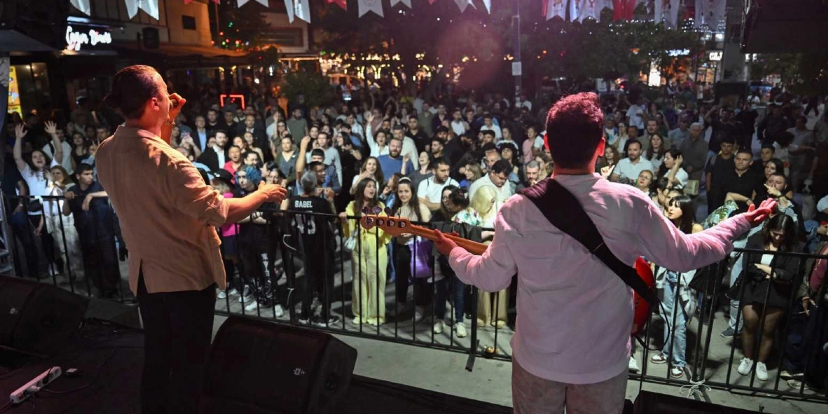 Açık havada düzenlenen bir konserde, sahneden kalabalığa doğru dönmüş iki müzisyen; solda mikrofonla şarkı söyleyen bir vokalist, sağda ise bas gitar çalan bir müzisyen yer alıyor. Sahne önündeki bariyerlerin arkasında, ellerini havaya kaldırmış veya müziğe eşlik eden coşkulu bir izleyici grubu bulunuyor. Gece vakti çekilen bu karede, arka planda dükkan ışıkları ve asılı süslemeler seçiliyor.