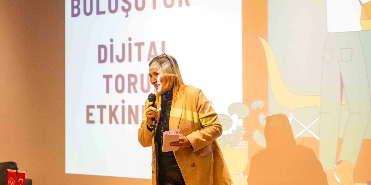 Sarı ceketli bir kadın, elinde mikrofonla sahnede konuşma yapıyor. Arkasındaki dev ekranda 