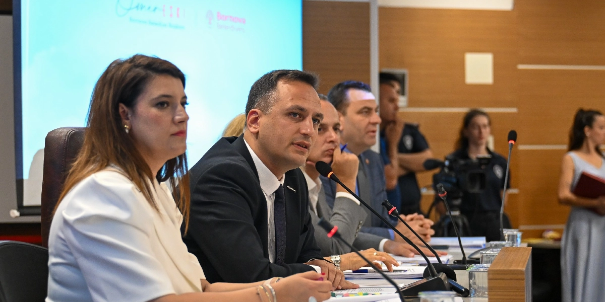Bornova Belediye Başkanı Mustafa İduğ ve beraberindeki heyetin katıldığı bir toplantıda, konuşmacılar masada mikrofonlar önünde otururken görülüyor. Arka planda Bornova Belediyesi logosunun yer aldığı bir projeksiyon ekranı bulunuyor.