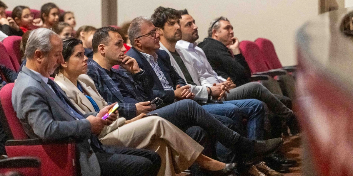 Konferans salonunda kırmızı koltuklarda yan yana oturan, dikkatle sahneye bakan bir grup yetişkin ve arka sıralarda oturan çocuklar. Ön plandaki katılımcılardan bazıları ellerinde telefon tutarken, bazıları ellerini çenesine dayamış veya kavuşturmuş şekilde sunumu takip ediyor.