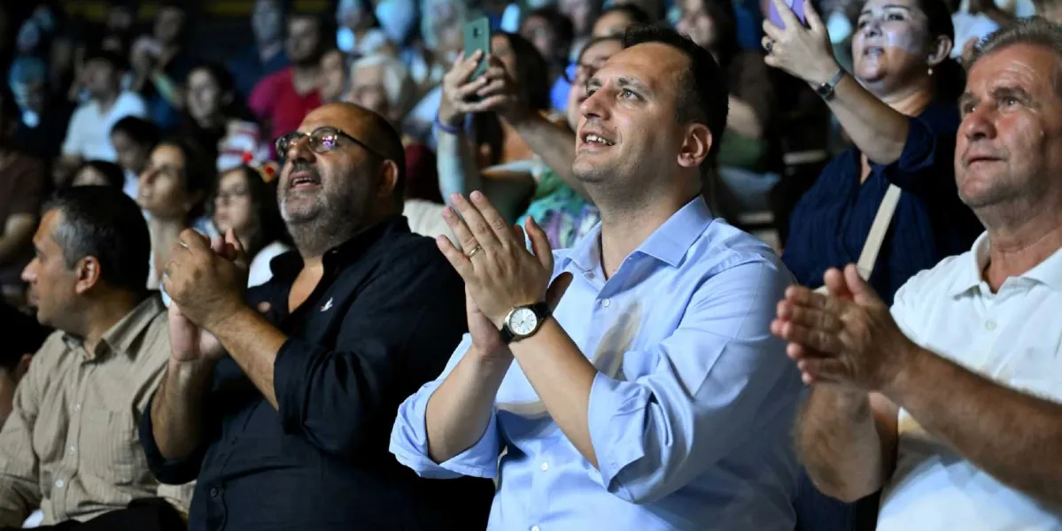 Kapalı bir mekanda, sahnedeki bir gösteriyi izleyen ve alkışlayan kalabalık bir grup insan. Ön planda, açık mavi gömlekli, kolunda saati olan bir erkek, yanında siyah gömlekli gözlüklü bir adam ve beyaz polo yaka tişörtlü başka bir erkekle birlikte coşkuyla alkışlıyor. Arka planda ise cep telefonlarıyla görüntü kaydeden veya sahneye odaklanmış çok sayıda izleyici bulunuyor.