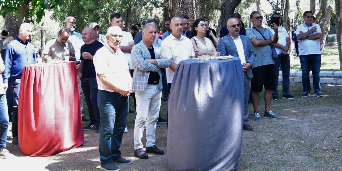 Açık havada, ağaçlık bir alanda düzenlenen anma etkinliğinde bir araya gelmiş, ayakta duran ve dikkatle dinleyen bir grup insan. Ön planda, üzerleri örtülü iki adet yüksek kokteyl masası bulunuyor; masaların üzerinde küçük ikramlar yer alıyor. Katılımcıların çoğu günlük kıyafetler içinde, bazıları kollarını kavuşturmuş, bazıları ise ellerinde telefonlarıyla çevreyi izliyor. Arka planda ise ağaçların gölgesinde bekleyen diğer katılımcılar görülüyor.