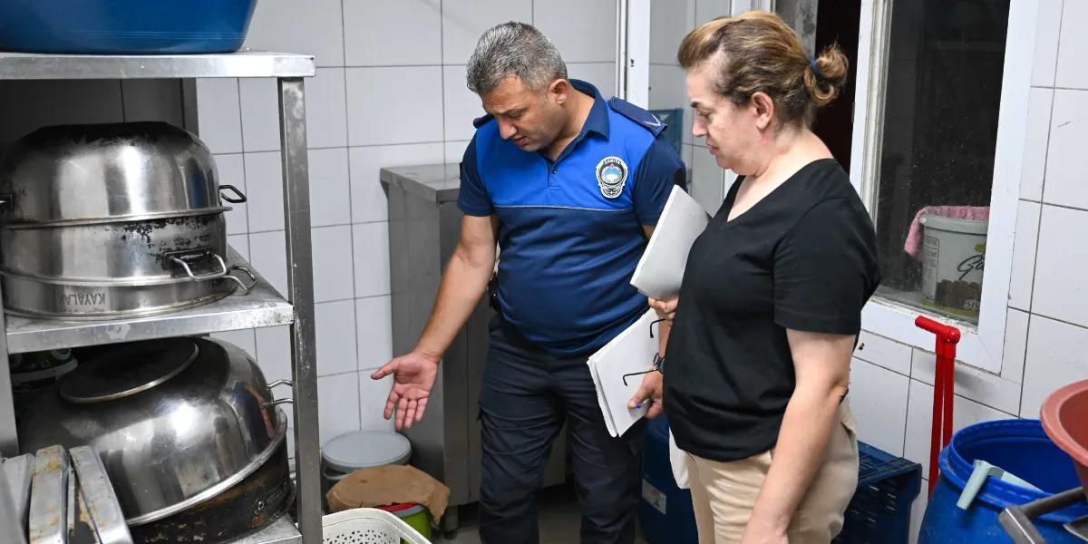 Zabıta üniformalı bir görevli, elinde dosyalarla bir fırın mutfağında inceleme yaparak bir noktayı işaret ediyor. Yanında duran kadın görevli ise elindeki belgelerle denetimi takip ediyor. Mutfak ortamında metal raflar üzerinde büyük pişirme kapları ve çeşitli ekipmanlar bulunuyor.