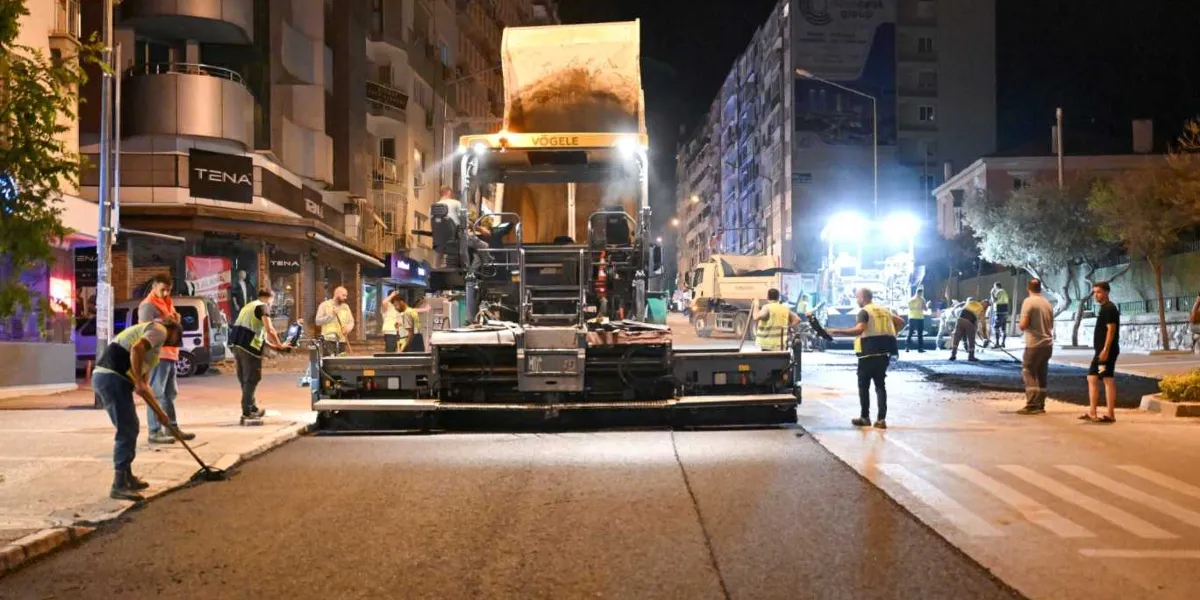 Gece vakti Mustafa Kemal Caddesi'nde devam eden yol yenileme çalışmaları kapsamında, sarı renkli büyük bir asfalt serme makinesi caddenin ortasında duruyor. İş güvenliği yelekleri giymiş bir grup işçi, makinenin çevresinde ve yol üzerinde asfalt serme ve düzenleme işlemlerini yürütüyor. Çevredeki binaların ışıkları ve iş makinelerinin güçlü aydınlatmalarıyla aydınlanan caddede, asfalt döküm süreci devam ediyor.