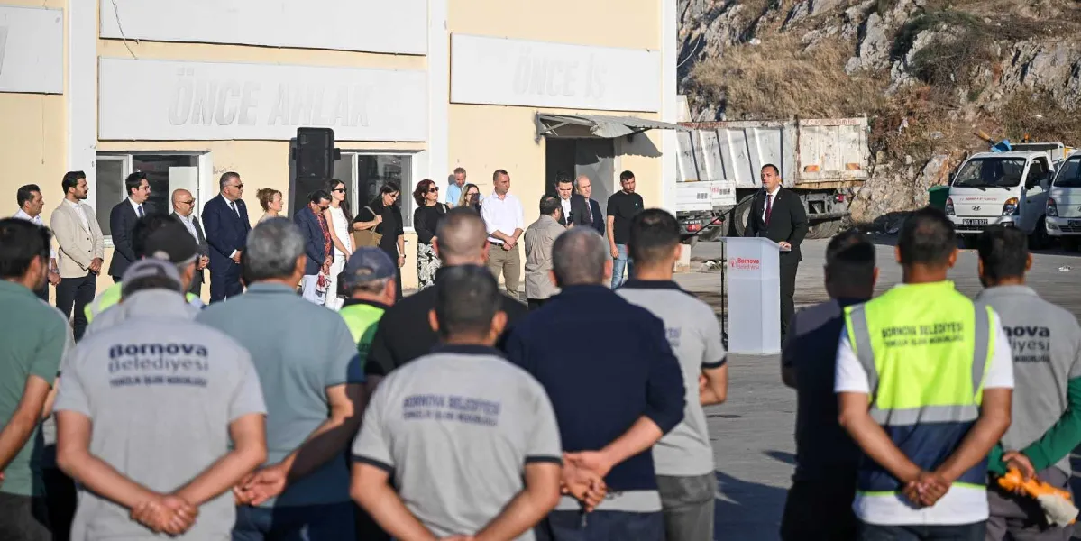 Bornova Belediye Başkanı Mustafa İduğ, üzerinde Bornova Belediyesi Temizlik İşleri Müdürlüğü yazan üniformalı işçilere ve bir grup katılımcıya açık havada hitap ediyor. Konuşma kürsüsünün arkasında, üzerinde 