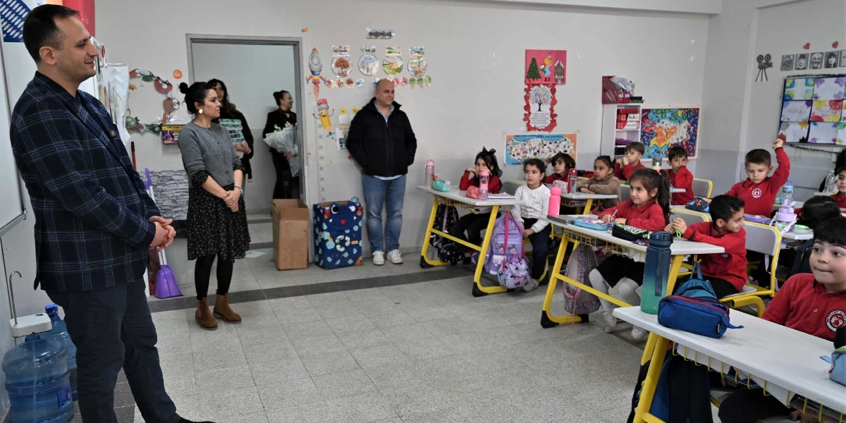 Koyu renkli ekose bir ceket giyen bir erkek, sınıfta sıralarda oturan ilkokul öğrencilerine hitap ediyor. Öğretmen olduğu düşünülen gri kazaklı bir kadın, sınıfın kapısı önünde duran diğer yetişkinlerle birlikte onu dinliyor. Kırmızı okul forması giymiş öğrenciler sıralarında oturarak dikkatle konuşmacıyı izliyor, bazıları parmak kaldırıyor. Sınıfın duvarları çocukların yaptığı resimler, haritalar ve eğitici görsellerle süslenmiş durumda.