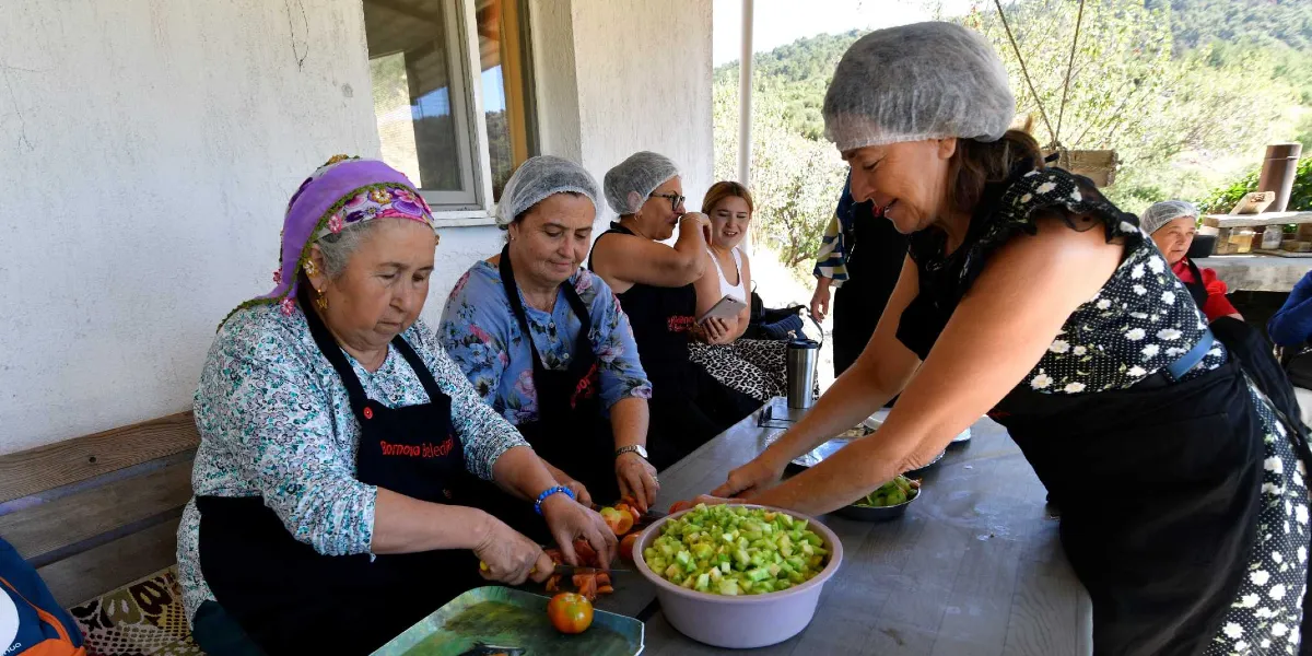 Açık havada, bir masa etrafında toplanmış, başlarında bone takan ve üzerinde Bornova Belediyesi yazılı siyah önlükler bulunan kadınlar sebze doğruyor. Ön planda bir kadın elindeki bıçakla domatesleri dilimlerken, yanında büyük bir kasede doğranmış yeşil sebzeler bulunuyor. Arka planda ise diğer kadınlar hazırlık yapmaya devam ediyor ve gülümseyerek birbirleriyle sohbet ediyor.