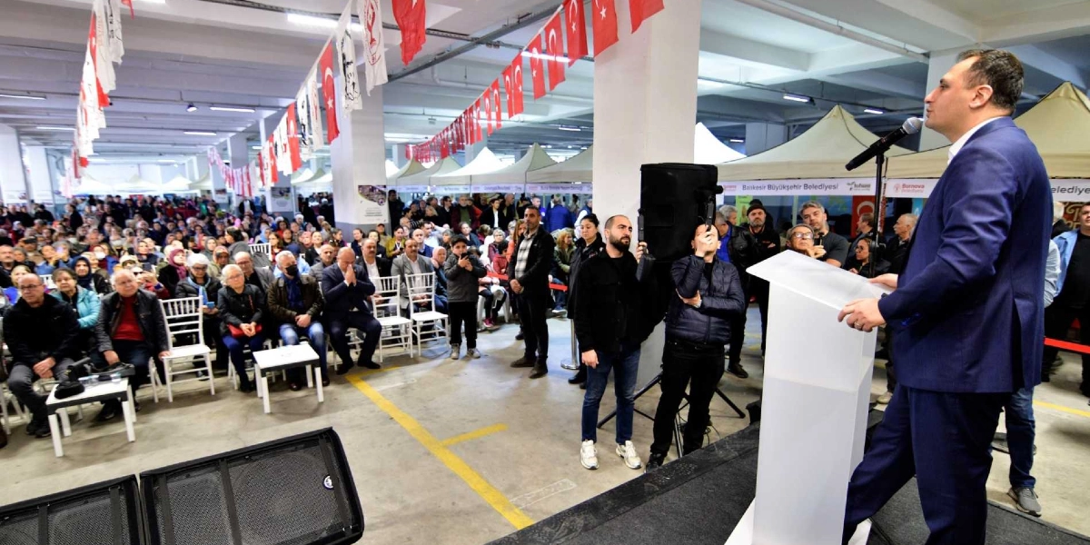 Kapalı bir alanda düzenlenen etkinlikte, kürsüde konuşma yapan lacivert takım elbiseli bir erkek ve onu dinleyen kalabalık bir izleyici grubu yer alıyor. Tavandan sarkan Türk bayrakları ve çeşitli flamalarla süslenmiş geniş alanda, izleyiciler sandalyelerde otururken, kürsünün hemen önünde çekim yapan kameramanlar bulunuyor. Arka planda ise stantların kurulduğu ve katılımcıların yoğun ilgi gösterdiği bir şenlik ortamı görülüyor.