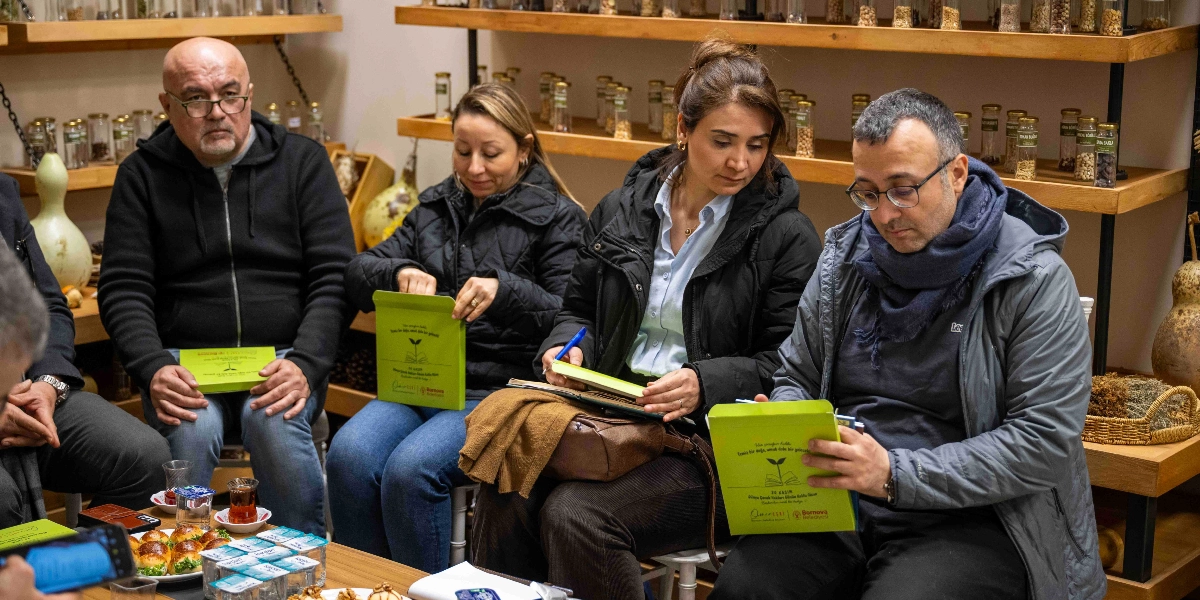 Kapalı bir mekanda, raflarında çok sayıda tohum kavanozu bulunan bir duvarın önünde oturan dört kişi, onarıcı tarım üzerine bir toplantı gerçekleştiriyor. Katılımcılar ellerinde Bornova Belediyesi logolu, üzerinde 