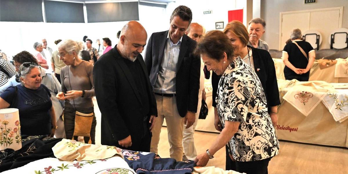 Atatürkçü Düşünce Derneği üyelerinin el emeği ürünlerini sergilediği bir etkinlikte, katılımcılar masaların üzerine serilmiş işlemeli örtüleri ve el işi ürünleri inceliyor. Ön planda, siyah desenli bir bluz giymiş bir kadın, masadaki bir kumaşı yakından incelerken yanında duran iki erkek ve diğer katılımcılarla sohbet ediyor. Arka planda ise el işi ürünlerin sergilendiği stantlar ve ürünleri inceleyen diğer ziyaretçiler yer alıyor.