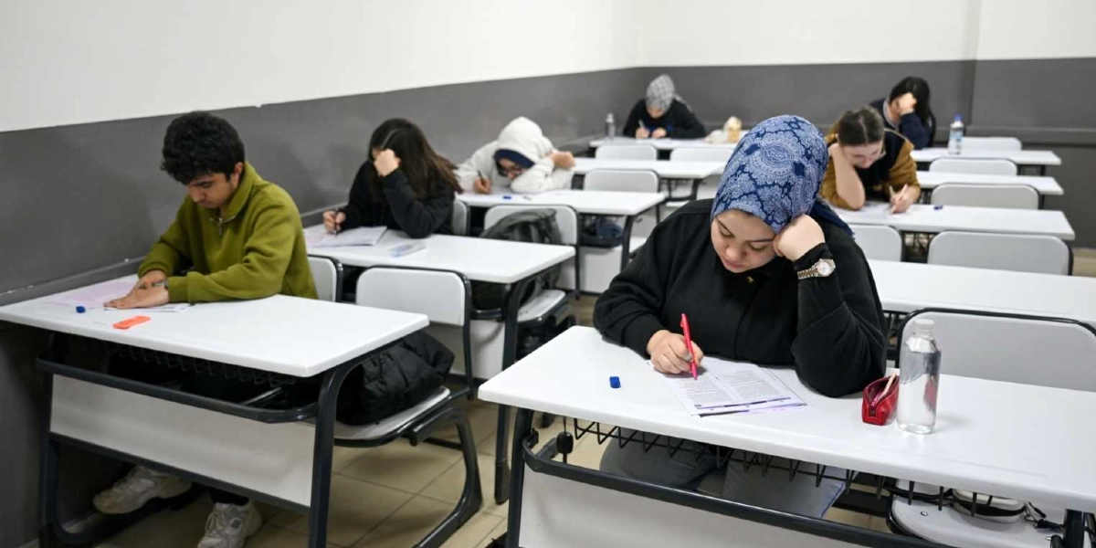 Sınıf ortamında tekli sıralarda oturan öğrenciler, önlerindeki sınav kağıtlarına odaklanmış bir şekilde deneme sınavına giriyor. Ön planda, başında mavi desenli bir eşarp olan ve elinde kırmızı bir kalemle sınav kağıdını dolduran bir öğrenci ile sol tarafında yeşil sweatshirt giymiş bir öğrenci dikkat çekiyor. Arka sıralarda ise farklı kıyafetler içindeki diğer öğrenciler sessizce sınavlarına devam ediyor. Masaların üzerinde su şişeleri ve kalemlikler bulunuyor.