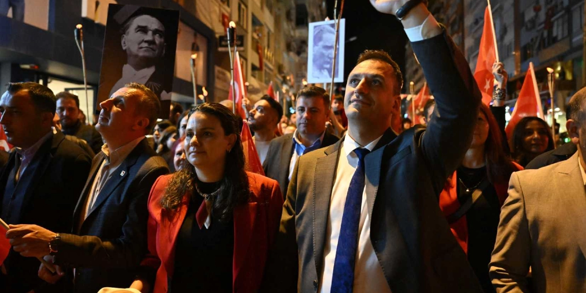 Gece saatlerinde düzenlenen bir Cumhuriyet Bayramı yürüyüşünde, takım elbiseli erkekler ve kırmızı ceketli kadınların yer aldığı kalabalık bir grup ellerinde Türk bayrakları ve Mustafa Kemal Atatürk posterleri taşıyor. Ön planda, koyu renk takım elbiseli ve mavi kravatlı bir erkek, elini havaya kaldırarak yürüyüşe öncülük ediyor. Çevredeki katılımcılar meşaleler eşliğinde ilerlerken, ortamda coşkulu bir atmosfer hakim.