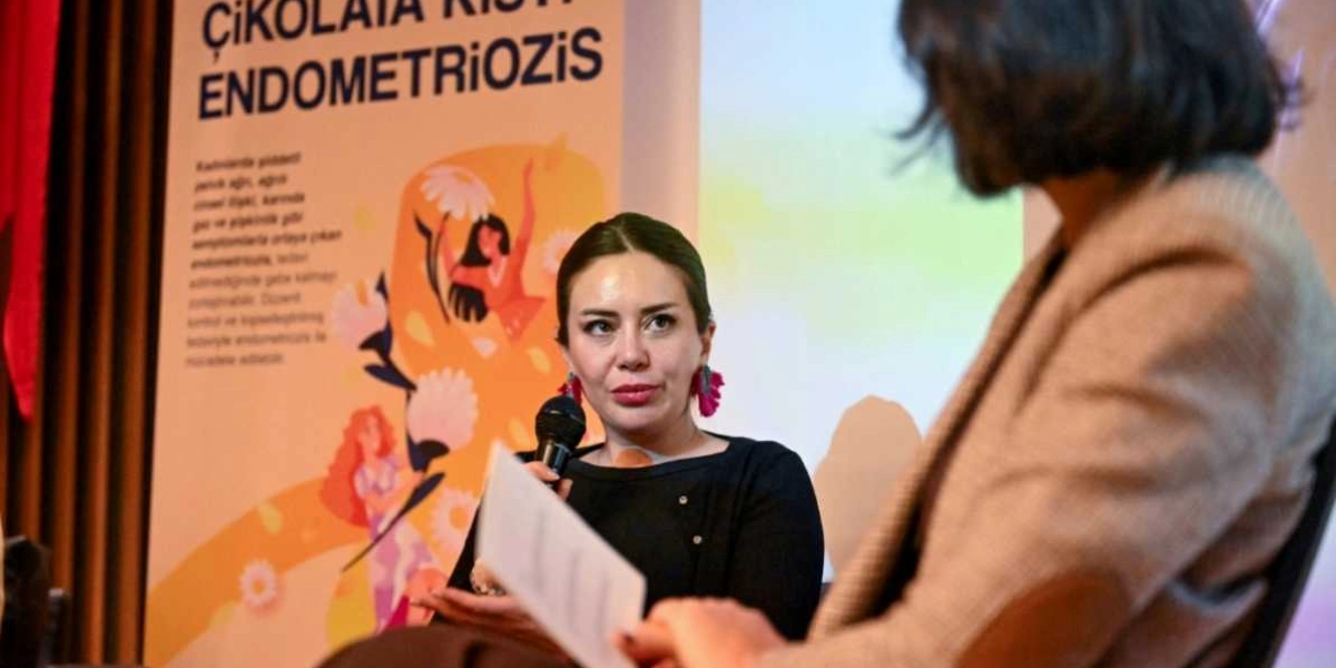 Siyah bluz giymiş, pembe sallantılı küpeler takan bir kadın, elinde mikrofonla konuşma yapıyor. Kadının arkasında üzerinde 
