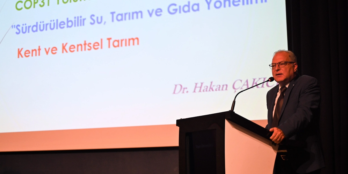 Kürsüde konuşma yapan gözlüklü ve takım elbiseli Dr. Hakan Çakıcı, arkasındaki projeksiyon ekranında 