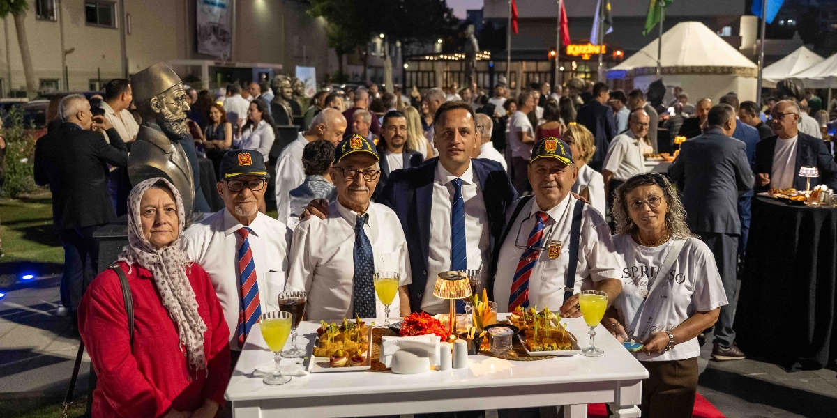 Açık havada düzenlenen bir akşam resepsiyonunda, beyaz bir kokteyl masasının etrafında bir araya gelmiş bir grup insan. Ön planda, üzerinde atıştırmalık tabakları ve içecekler bulunan masanın arkasında; beyaz gömlekli, lacivert kravatlı ve üzerinde 