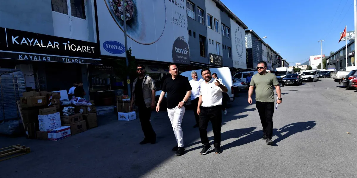 Güneşli bir günde Fincancılar Çarşısı'nda yürüyen Başkan Eşki ve beraberindeki heyet. Başkan Eşki beyaz gömlek ve siyah pantolon giymiş, eliyle bir yeri işaret ederek yanındakilerle sohbet ediyor. Çevredeki dükkanların tabelalarında Kayalar Ticaret, Bonna ve Yontaş gibi markalar yer alıyor; dükkan önlerinde ise istiflenmiş koli ve ürünler bulunuyor.