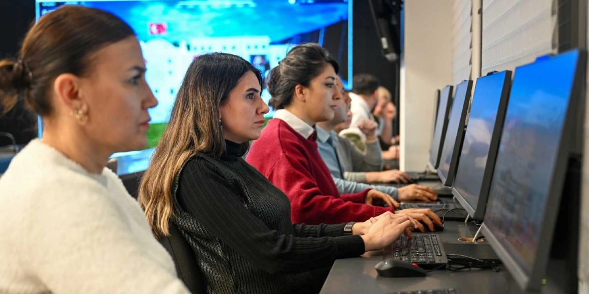 Modern bir ofis ortamında yan yana dizilmiş bilgisayarların başında çalışan üç kadın personel, odaklanmış bir şekilde ekranlarına bakarak klavye ve fare kullanıyor. Arka planda ise büyük bir dijital ekran üzerinde Bornova Belediyesi binasının görseli yer alıyor.
