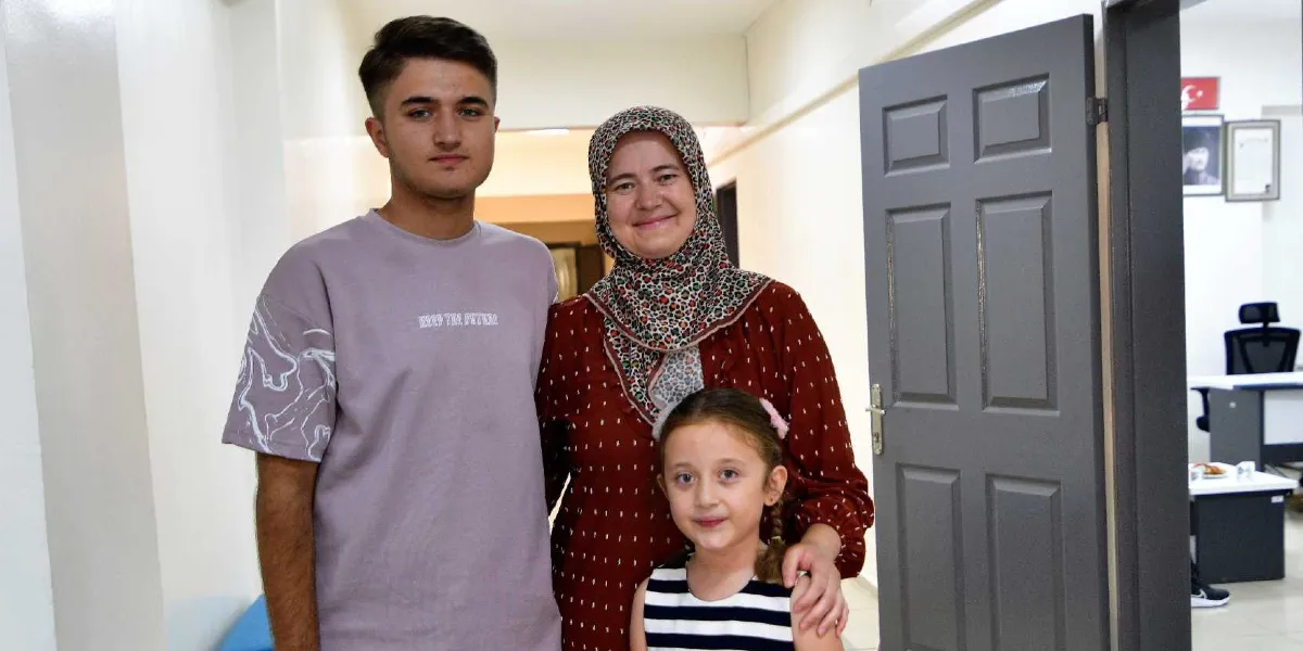 İç mekanda yan yana duran genç bir erkek, başörtülü bir kadın ve küçük bir kız çocuğu. Genç erkek üzerinde 