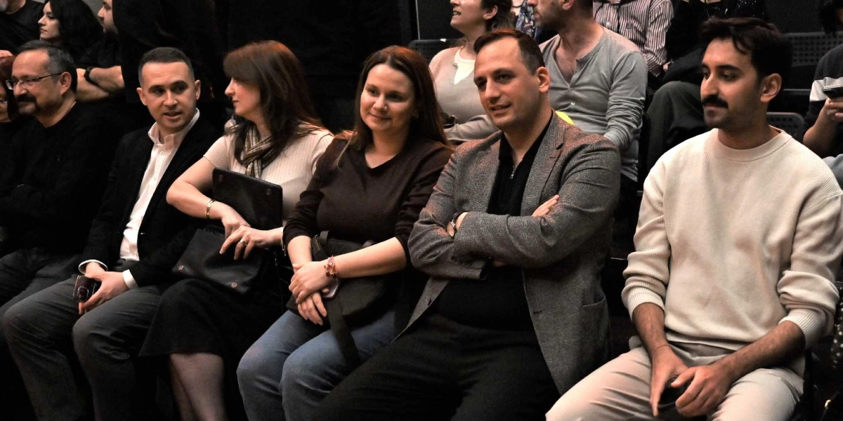 Kapalı bir salonda, tiyatro veya konferans izlemek üzere yan yana oturmuş bir grup insan. Ön planda, bej rengi kazaklı bıyıklı bir genç adam, yanında gri ceketli ve kollarını kavuşturmuş bir erkek, onun yanında koyu renk bluzlu bir kadın, yanında açık renk bluzlu ve elinde siyah bir çanta tutan bir kadın ile siyah ceketli bir erkek yer alıyor. İzleyiciler dikkatle sahneye doğru bakıyor ve genel olarak yarı resmi bir kıyafet tarzı sergiliyorlar.