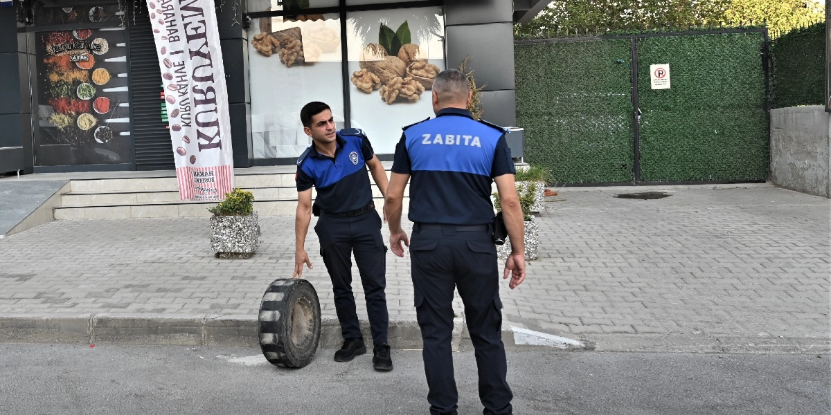 Üniformalı iki zabıta memuru, bir kuruyemiş dükkanının önündeki kaldırımda duruyor. Memurlardan biri, kaldırım işgali yapan büyük bir lastiği eliyle işaret ederek diğeriyle konuşuyor. Arka planda dükkanın camında kuruyemiş görselleri ve üzerinde 