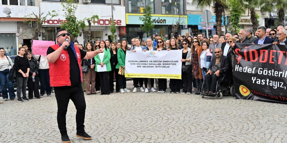 Bornova'da bir meydanda toplanan kalabalık grup, güvenli eğitim talebiyle basın açıklaması yapıyor. Ön planda, üzerinde kırmızı yelek olan bir kişi elinde mikrofonla konuşma yaparken, arkasında ellerinde 