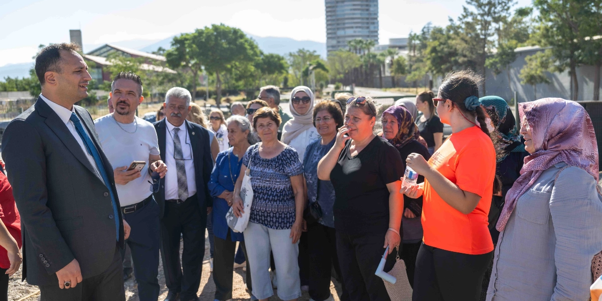 Bornova'da düzenlenen bir etkinlikte, takım elbiseli bir yetkili, açık havada toplanmış bir grup vatandaşa hitap ediyor. Katılımcılar arasında farklı yaş gruplarından kadınlar ve erkekler bulunuyor; grup dikkatle konuşmacıyı dinliyor. Arka planda ağaçlar, geniş bir alan ve uzakta yüksek bir bina görülüyor.