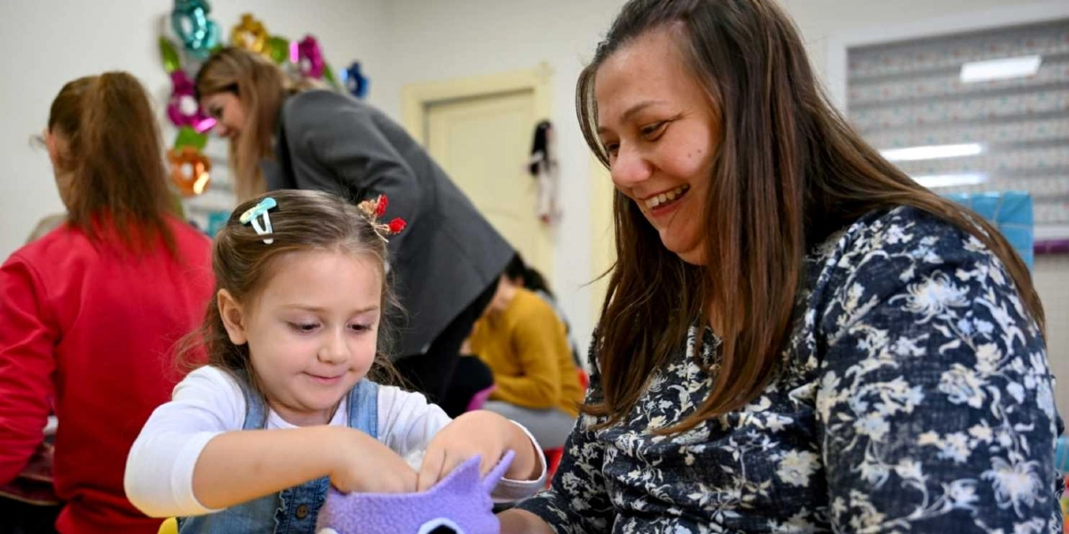 İç mekanda, çiçek desenli lacivert bir bluz giyen güler yüzlü bir kadın, yanında oturan küçük kız çocuğunun mor renkli bir saksıya toprak doldurmasına yardım ediyor. Arka planda ise başka yetişkinler ve çocuklar etkinlik masasında oturuyor.