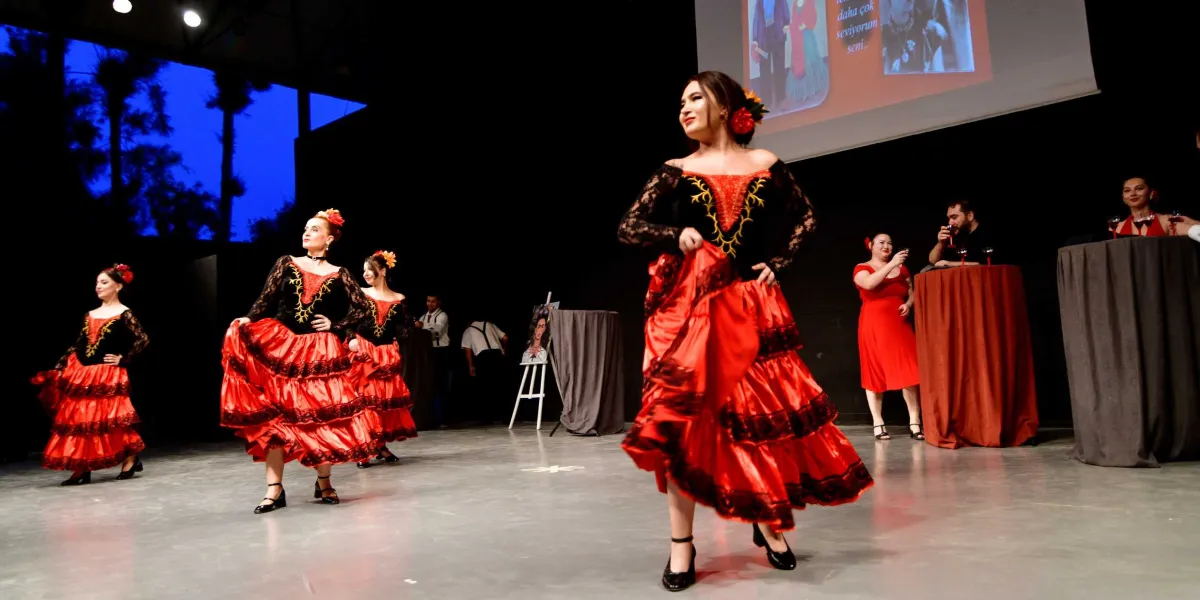 Siyah dantel detaylı üstleri ve kırmızı kat kat etekleriyle geleneksel İspanyol dansı yapan dört kadın dansçı, sahnede performans sergiliyor. Dansçıların saçlarında kırmızı çiçekler bulunuyor. Sahnenin arka planında, üzerinde Frida Kahlo'nun görselleri ve metinler bulunan büyük bir projeksiyon ekranı yer alıyor. Arka tarafta, kırmızı örtülü masaların başında duran ve içeceklerini yudumlayan diğer katılımcılar görülüyor.