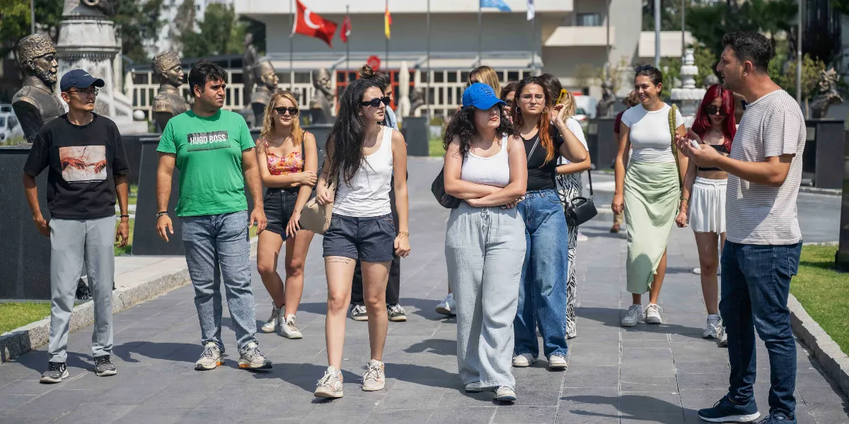 Abide-i Hürriyet Meydanı'nda, tarihi büstlerin ve Türk bayrağının bulunduğu açık alanda yürüyen genç bir grup ve onlara rehberlik eden bir erkek. Grup üyeleri gündelik kıyafetler içinde, güneşli bir havada meydandaki taş yolda ilerlerken rehber, elindeki bir nesneyle onlara açıklama yapıyor.