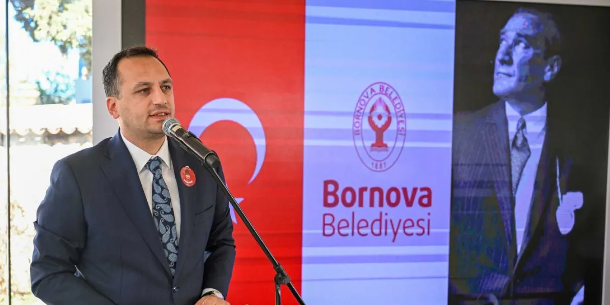 Koyu renk takım elbiseli ve desenli kravatlı bir erkek, önündeki mikrofona konuşma yapıyor. Konuşmacının yakasında Atatürk figürlü bir rozet bulunuyor. Arka planda ise üzerinde Bornova Belediyesi logosu ve yazısı bulunan, Türk bayrağı motifli bir ekran ve sağ tarafta Mustafa Kemal Atatürk'ün siyah beyaz bir portresi yer alıyor.