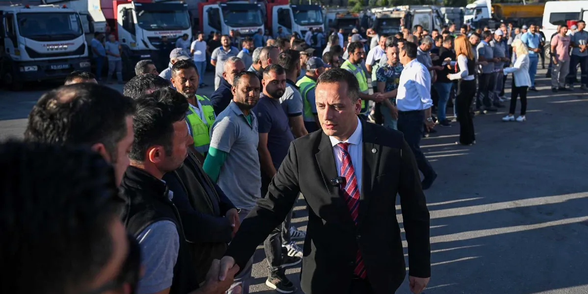 Siyah takım elbiseli ve kırmızı çizgili kravatlı bir erkek, açık havada bir grup işçi ve sendika temsilcisiyle tokalaşarak ilerliyor. Arka planda belediyeye ait olduğu anlaşılan çok sayıda çöp kamyonu ve onları izleyen kalabalık bir grup insan bulunuyor.