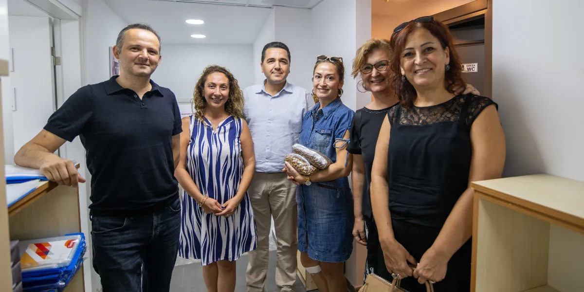 İç mekan bir koridorda yan yana duran, gülümseyerek kameraya bakan altı kişilik bir grup. En solda siyah polo yaka tişörtlü bir erkek, yanında beyaz üzerine mavi desenli kolsuz bir elbise giyen kadın, ortada açık renk gömlekli bir erkek, yanında elinde iki paket kuru mama tutan kot elbiseli bir kadın, onun yanında gözlüklü bir kadın ve en sağda siyah kolsuz bir bluz giyen kadın yer alıyor. Grup, bir çalışma ortamında bir araya gelmiş görünüyor.