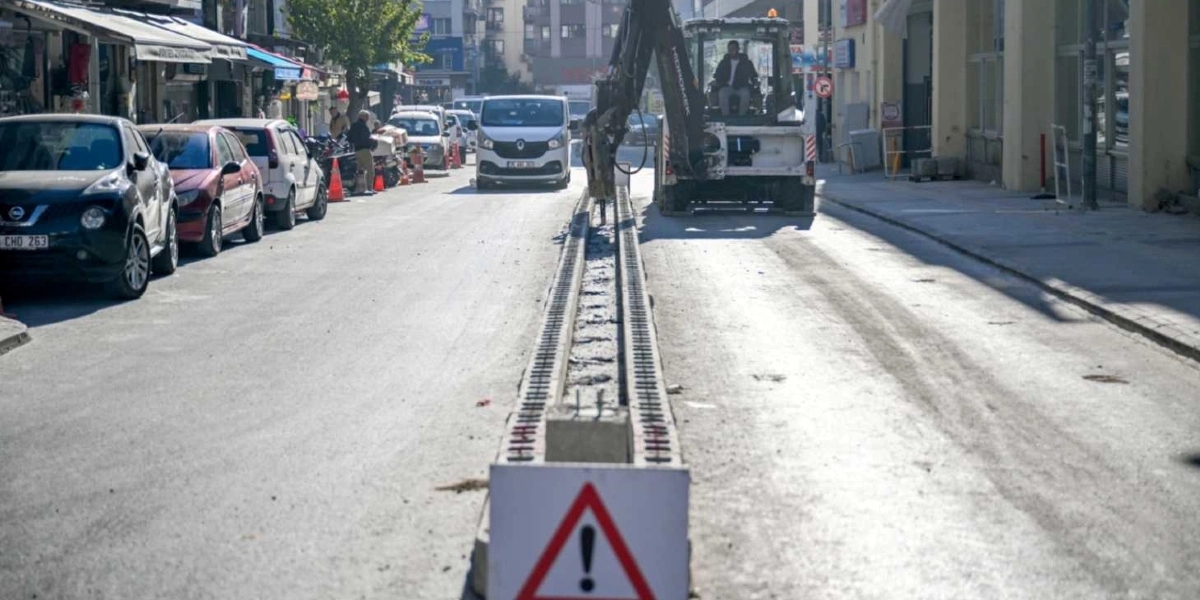 Şehir merkezindeki bir caddede, yolun ortasına yerleştirilmiş ray hattı üzerinde çalışan bir iş makinesi ve önünde üzerinde ünlem işareti bulunan bir uyarı levhası yer alıyor. Caddenin kenarlarında park halindeki araçlar ve dükkanlar görülürken, trafik akışı kontrollü bir şekilde devam ediyor.