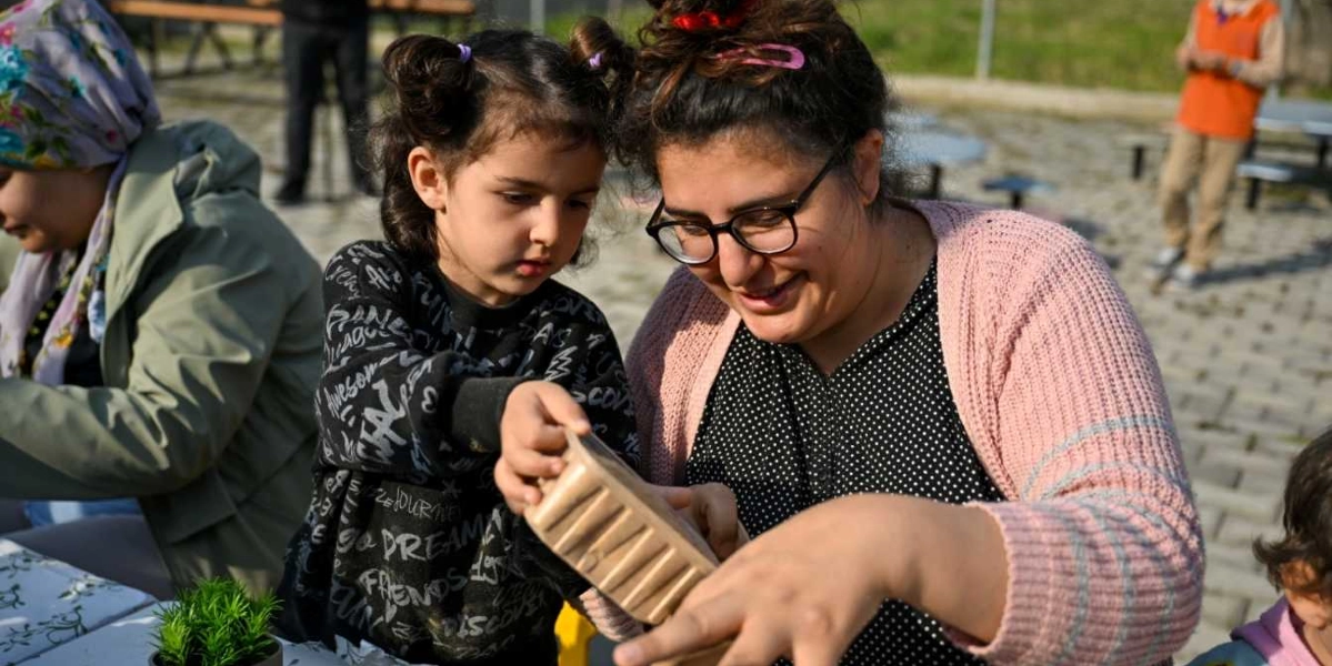 Açık havada düzenlenen bir atölye çalışmasında, gözlüklü bir kadın ve yanındaki küçük kız çocuğu, ellerindeki plastik bir kabı inceleyerek çim adam yapımıyla ilgileniyorlar. Arka planda başka katılımcıların da bulunduğu, parke taşlı bir alanda gerçekleşen etkinlikte, masanın üzerinde küçük bir saksı bitkisi yer alıyor.