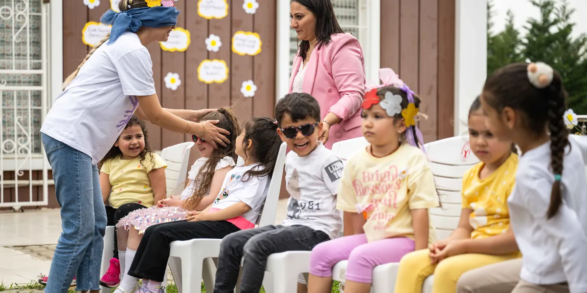 Gözleri mavi bir bantla kapatılmış bir kadın, bir anaokulu bahçesinde sandalyelerde oturan çocukların arasında dolaşarak onlara dokunuyor. Çocukların bir kısmı güneş gözlüğü takıyor ve başlarında çiçekli taçlar bulunuyor. Arka planda pembe ceketli bir kadın onları izliyor, kahverengi duvarın üzerinde ise papatya süslemeleri yer alıyor.