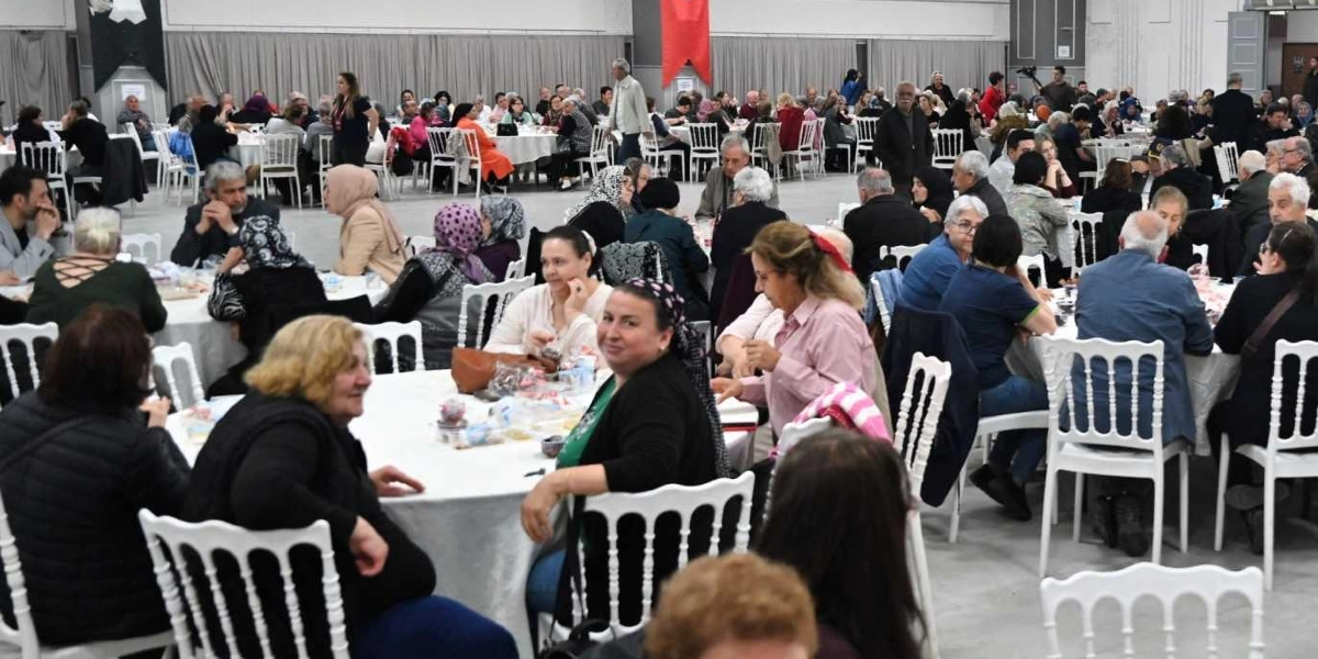 Geniş ve aydınlık bir salonda, beyaz örtülü yuvarlak masaların etrafında toplanmış çok sayıda kişinin katıldığı bir dayanışma yemeği. Farklı yaş gruplarından kadın ve erkeklerin bir arada oturduğu, sohbet ettiği ve yemek yediği kalabalık bir ortamda, ön planda yer alan masadaki katılımcılar kameraya bakıyor. Arka planda ise benzer şekilde düzenlenmiş çok sayıda masa ve katılımcı grubu görülüyor.