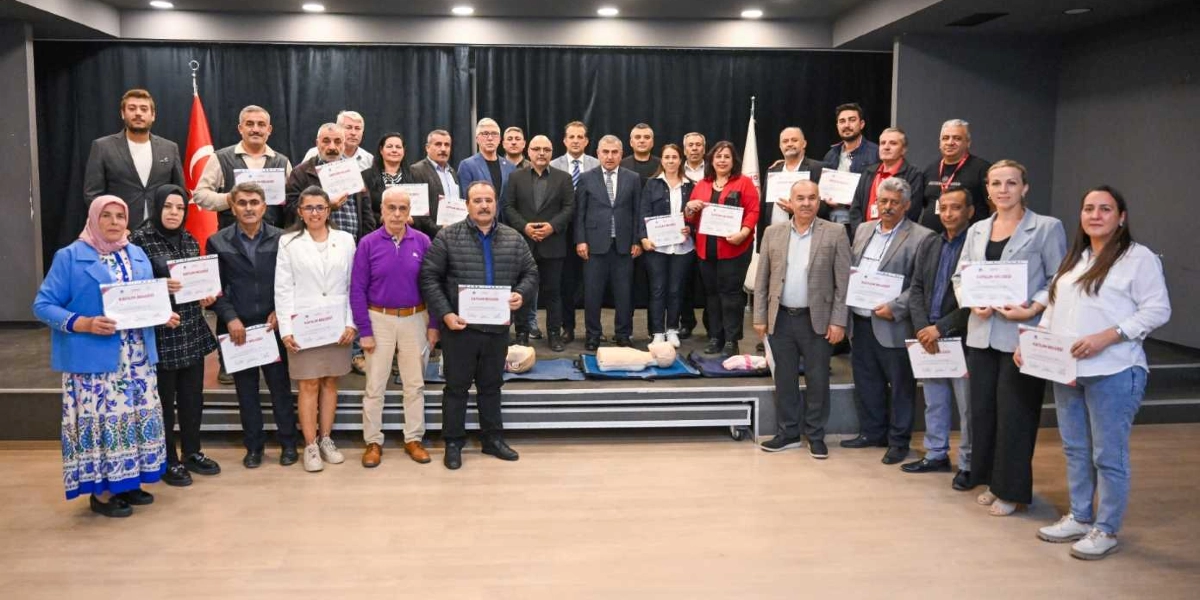 Kapalı bir salonda, ellerinde katılım belgeleriyle poz veren, aralarında Bornova muhtarlarının da bulunduğu kalabalık bir grup. Grubun önünde, yerde ilk yardım eğitiminde kullanılan iki adet cansız manken duruyor. Katılımcıların bir kısmı resmi kıyafetler, bir kısmı ise günlük kıyafetler giymiş durumda; arka planda ise Türk bayrağı ve Bornova Belediyesi'ne ait olduğu anlaşılan bir flama yer alıyor.