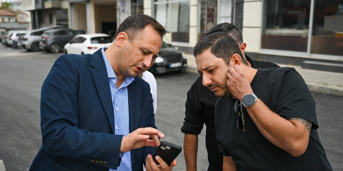 Lacivert ceketli ve mavi kareli gömlekli bir adam, elindeki akıllı telefondan bir şeyler göstererek yanındaki siyah tişörtlü iki kişiye açıklama yapıyor. Üç kişi, asfaltlanmış bir sokakta, park halindeki araçların önünde durarak telefondaki içeriği dikkatle inceliyor.