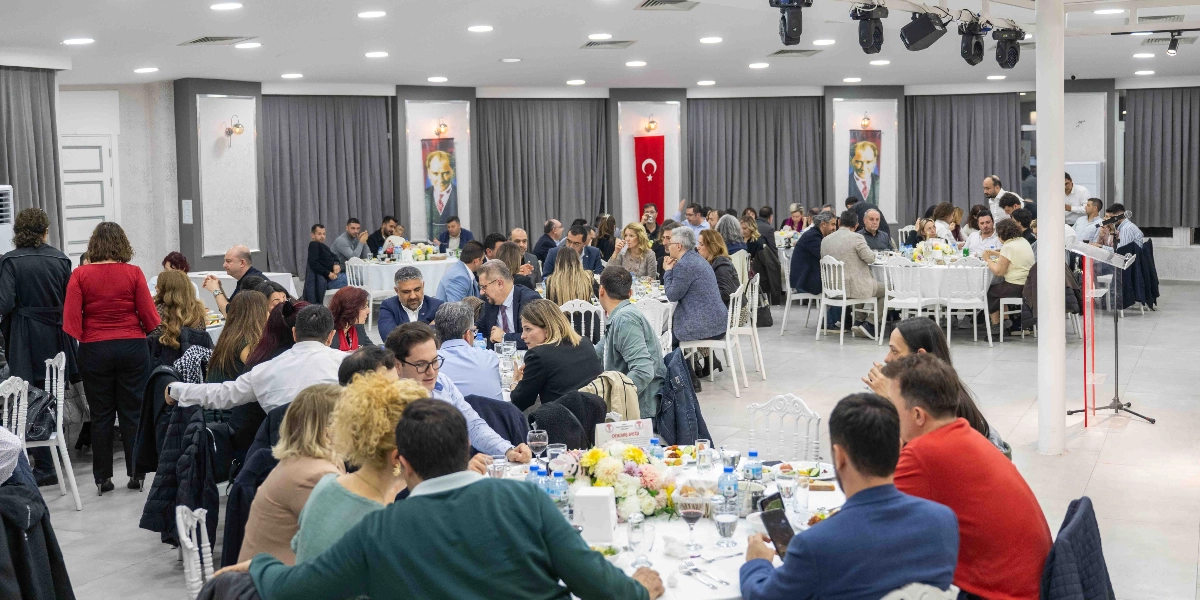 Geniş ve aydınlık bir salonda, yuvarlak masalar etrafında toplanmış çok sayıda kişinin katıldığı bir yemekli toplantı. Katılımcılar şık kıyafetlerle masalarda oturarak sohbet ediyor ve yemek yiyor. Salonun arka duvarında gri perdelerin önünde asılı olan Atatürk posterleri ve Türk bayrağı dikkat çekiyor. Tavanda spot ışıklar bulunurken, sağ tarafta bir kürsü ve arka planda ayakta duran veya masalarda oturan kalabalık bir davetli grubu yer alıyor.