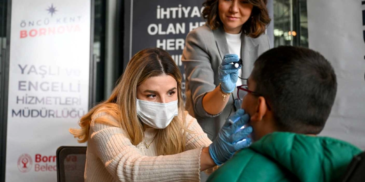 Maskeli bir diş hekimi, bir öğrencinin dişlerini muayene ederken arkasında duran bir başka uzman da elindeki ışıklı cihazla ağız içine bakıyor. Arka planda Bornova Belediyesi'ne ait 